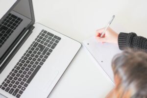 Seen from above, a person writes in a notebook in front of a laptop